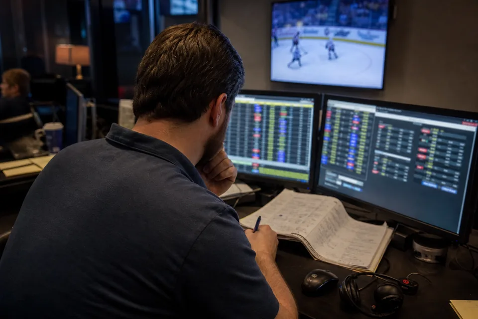 Odds trader at a sportsbook desk watching live price feeds across several screens during an evening game